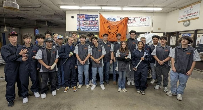 A large group of male and female teens poses for the camera in grey work uniforms while some in the back hold up an orange Bulldog flag and one holds a large wrench.