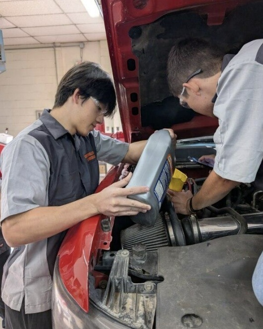 Two male teens work together to pour oil into a vehicle.