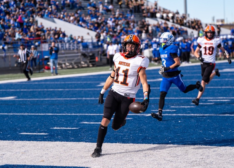 A teen boy in a white, orange and black football uniform runs off a blue football field into a white end zone with the ball in his left hand.
