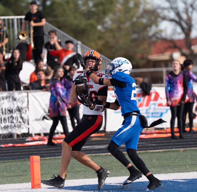 A teen boy in a white, orange and black football uniform catches a football just inside the orange pylon in front of an opponent in blue.