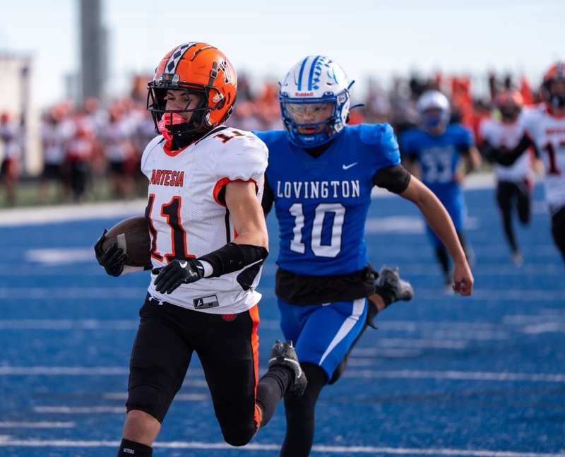 A teen boy in a white, orange and black football uniform runs ahead of an opponent in blue with the football tucked in his right elbow.