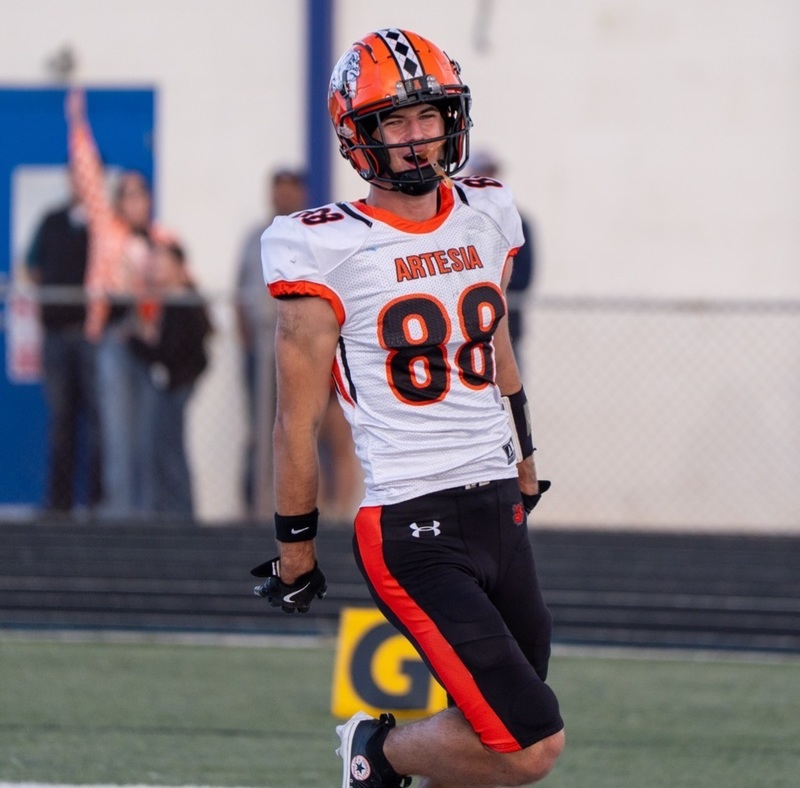 A teen boy in a white, orange and black football uniform smiles as he runs back through the end zone after catching a touchdown pass.