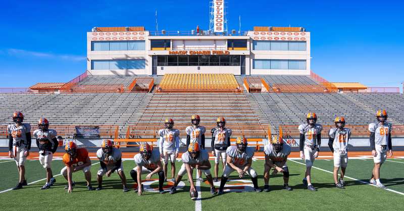 A group of boys in grey jerseys and orange helmets stands in an offensive formation on a football field.