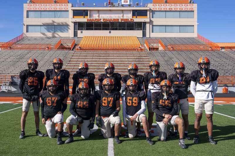 A group of boys in black jerseys and orange helmets poses for a photo on a football field.