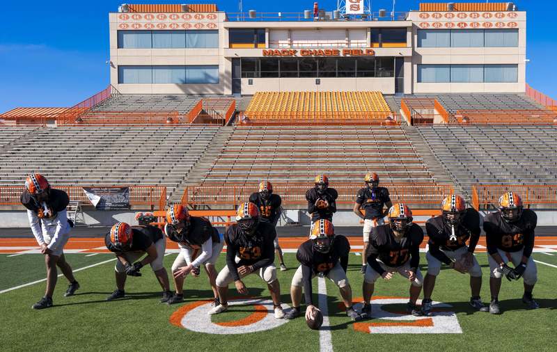 A group of boys in black jerseys and orange helmets poses in an offensive formation on a football field.