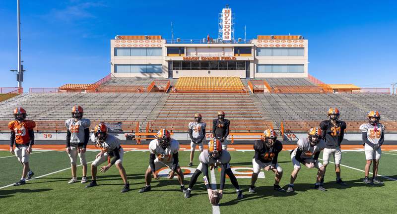 A group of boys in practice football jerseys poses in a punting formation on a football field.
