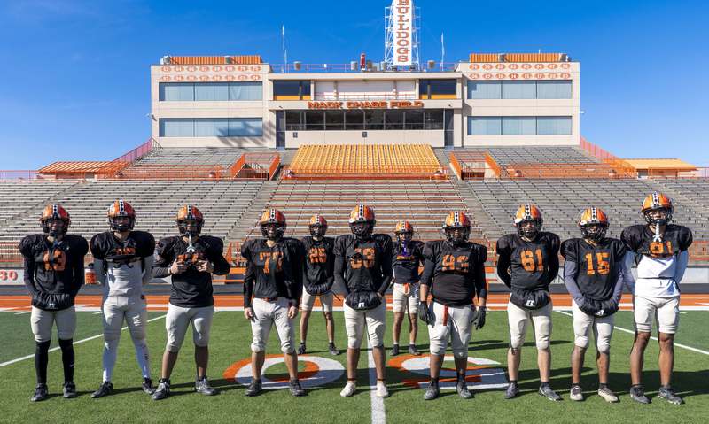 A group of boys in black jerseys and orange helmets poses for a photo in a punt return formation.