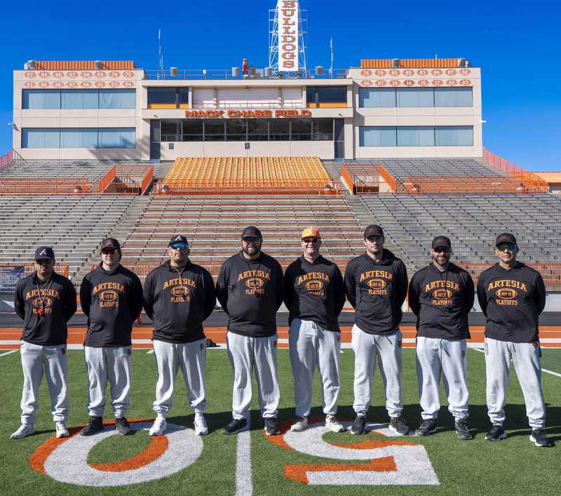 A group of eight men in black hoodies and grey sweatpants poses for a photo on a football field.