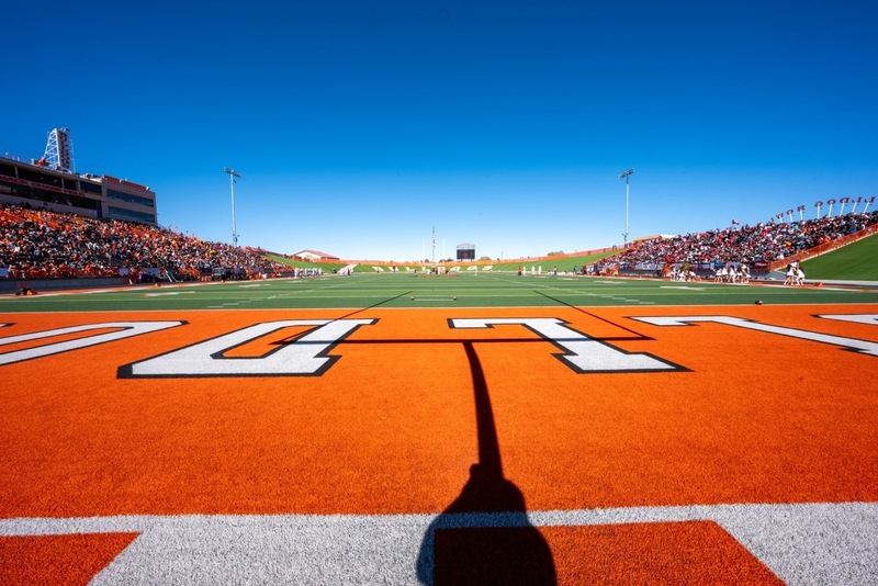 A view of a football field with an orange end zone, with stands on either side packed with fans.