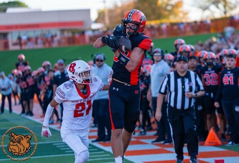 A teen boy in a black and orange football uniform twists to the side to receive a football in front of an opponent in white and red.
