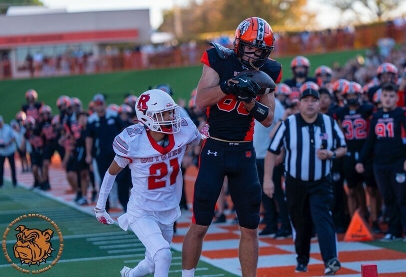 A teen boy in a black and orange football uniform brings in a football in front of an opponent in red and white.