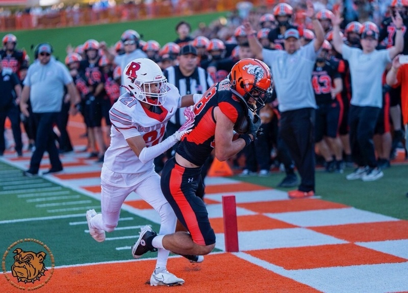A teen boy in a black and orange football uniform comes down in an orange end zone in front of an opponent in white and red as coaches and teammates cheer and signal touchdown in the background.