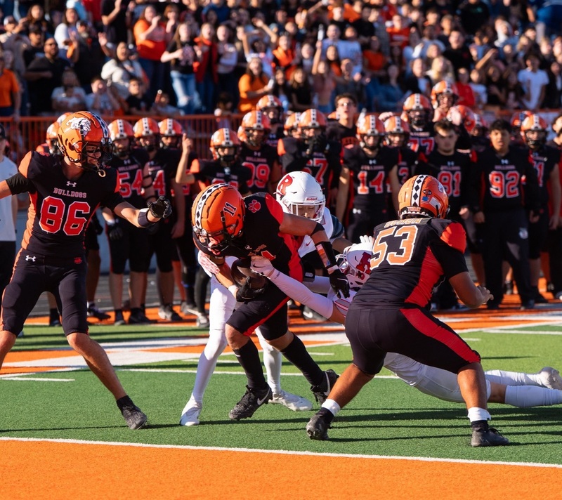 A teen boy in a black and orange football uniform pushes toward the goal line as two opponents in white and red hang from his body.