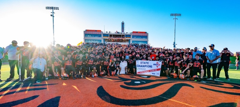 A high school football team in black and orange football uniforms pose in front of a large grandstand with a blue trophy, banner and bracket.