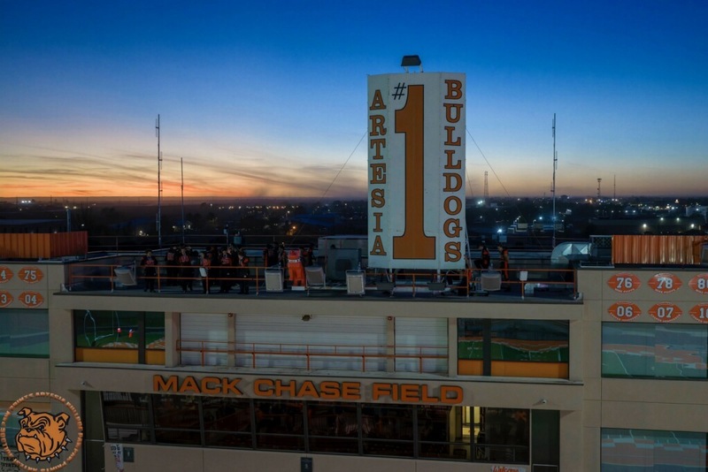 A press box is seen from the air with a blue and orange sunset behind it. Teen boys in black and orange football uniforms stand atop it next to a large white season reading "Artesia Bulldogs #1" in orange.