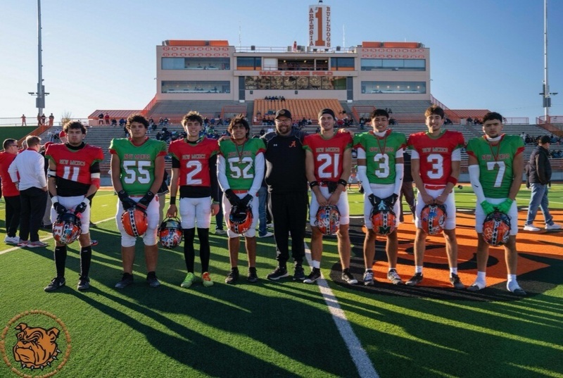 A group of teen boys, four in red football jerseys and four in green, stand in a line holding their orange helmets. A male coach poses between them.