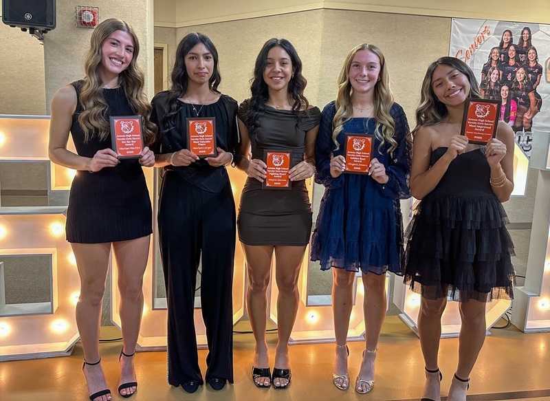 Five teen girls, four in dresses and one in a blouse and slacks, pose for a photo holding orange plaques.