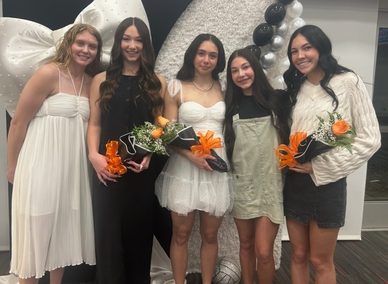 Five teen girls in dresses pose for a photo. Three are holding bouquets of orange roses.