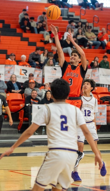 A teen boy in an orange basketball uniform puts up a shot between two defenders in white.