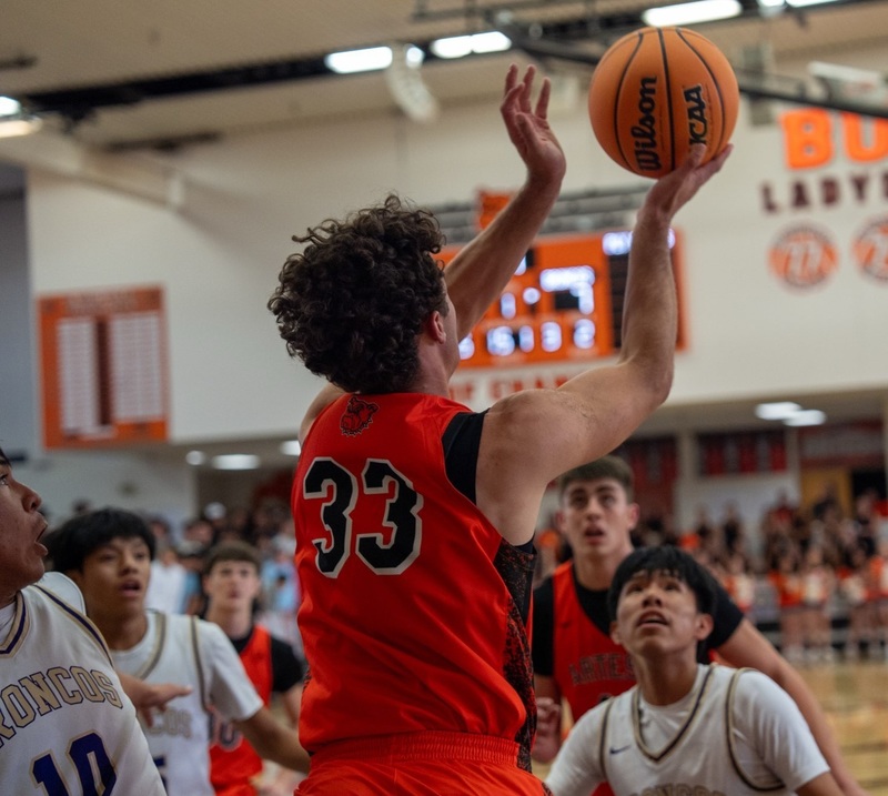 The back of a teen boy in an orange basketball uniform is seen as he prepares to lay the basketball off the backboard.