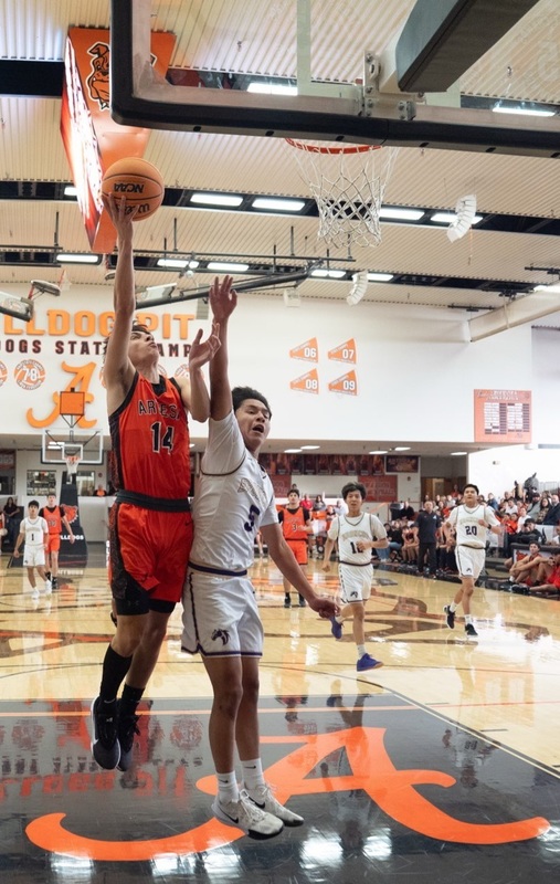 A teen boy in an orange basketball uniform prepares to lay the ball off the glass ahead of a defender in white.