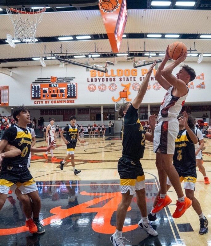 A teen boy in a white and orange basketball uniform jumps in the air as he prepares to release the basketball over a defender in black and gold.