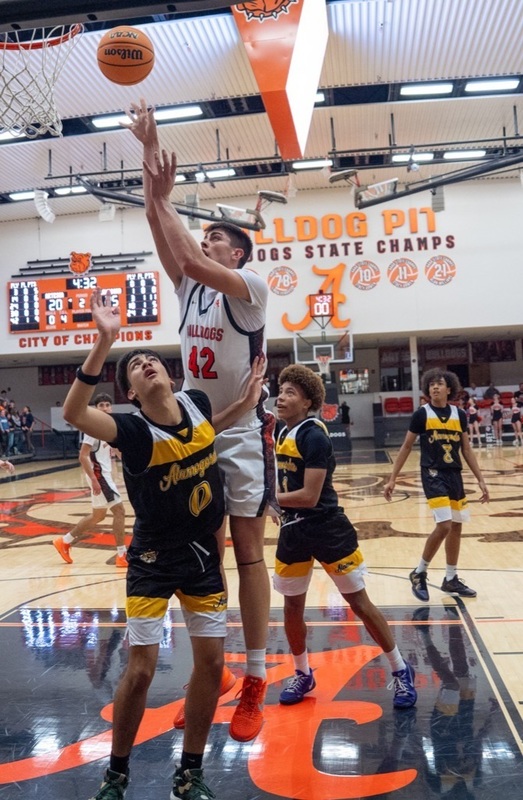 A teen boy in a white and orange basketball uniform puts the ball off the glass in mid-air over a defender in black and gold.