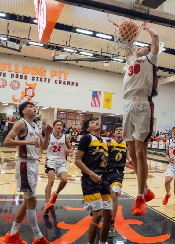 A teen boy in a white and orange basketball uniform comes off the rim after dunking the ball as two teammates and two defenders in black and gold look on.