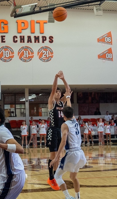 A teen boy in a black and orange basketball uniform shoots the ball high into the air while jumping as a defender in white and blue raises an arm in front of him.