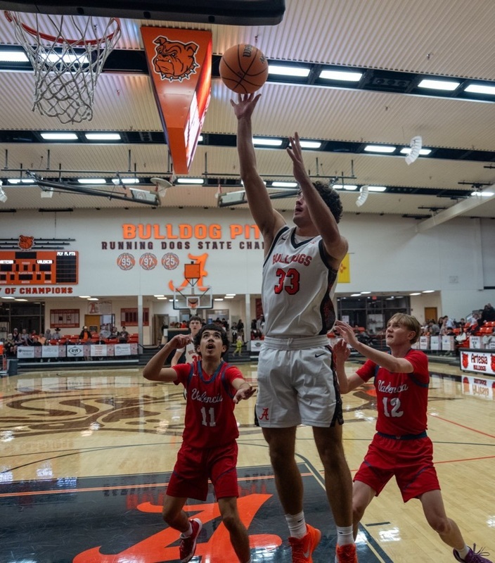 A teen boy in a white and orange basketball uniform releases the ball from his fingertips as he jumps toward the basket between to defenders in red.