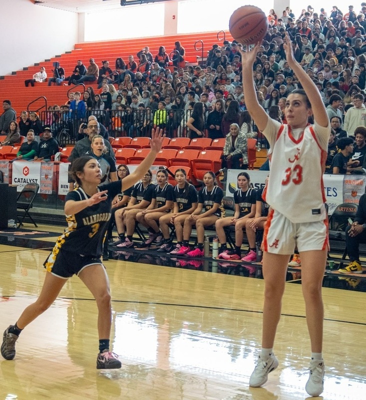 A teen girl in a white and orange basketball uniform prepares to release the ball ahead of an opponent in black and gold as a large crowd looks on.