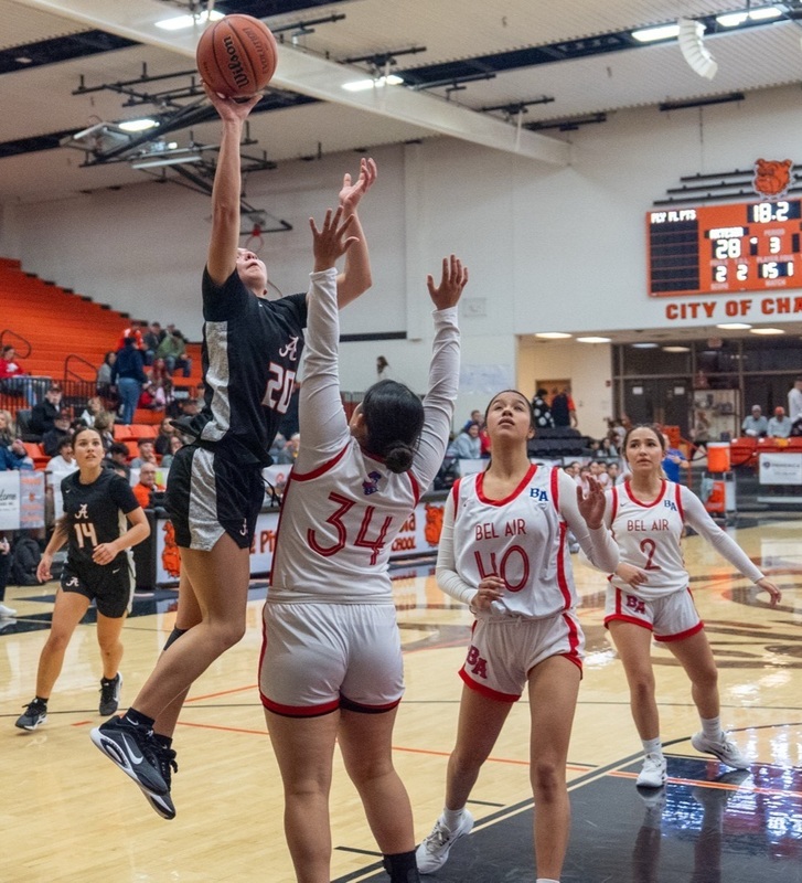A teen girl in a black and orange basketball uniform goes airborne to shoot over an opponent in white and red.