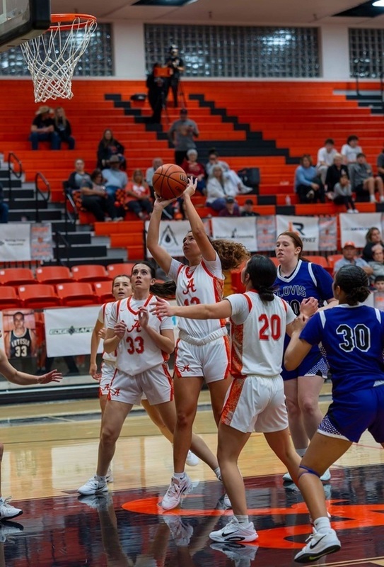 A teen girl in a white and orange basketball uniform prepares to shoot the ball between a pair of teammates and two opponents in purple.