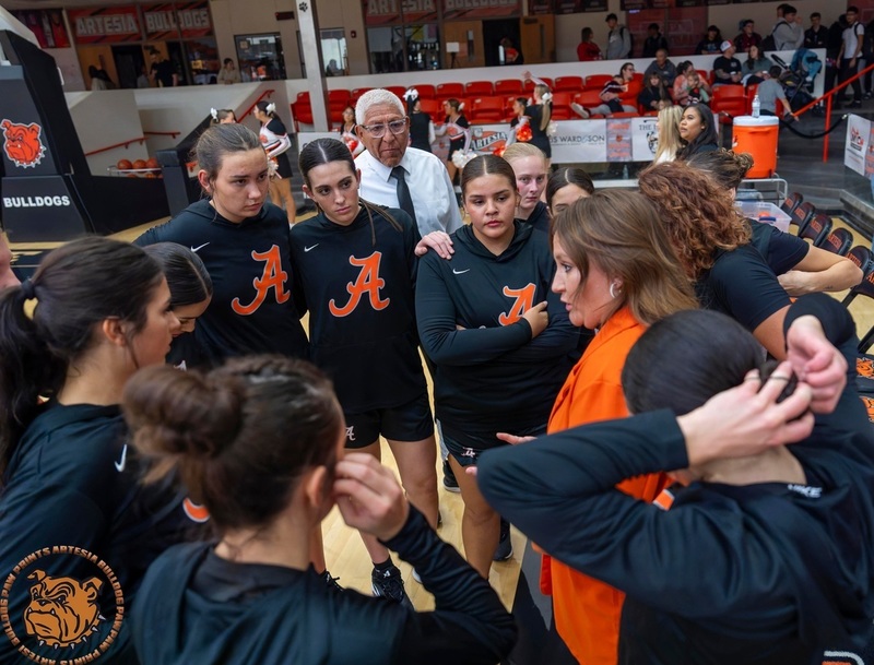 A group of teen girls in black warm-up shirts with orange A's on the front listen as a female coach in orange speaks to them in the huddle.