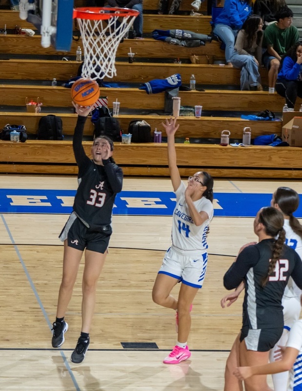 A teen girl in a black and white basketball uniform jumps to shoot with her right hand past an opponent in white and blue.