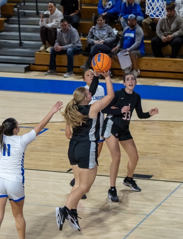 A teen girl in a black and white basketball uniform is seen from behind as she lunges toward the basket with the ball in her right hand.