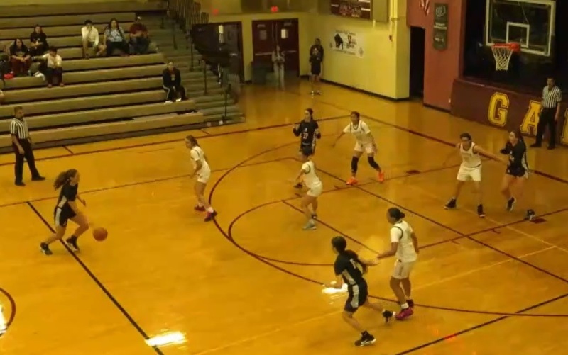 In a wide shot in a dimly lit gym, a teen girl in a black and white basketball uniform is seen dribbling toward the top of the key as teammates and opponents in white watch.
