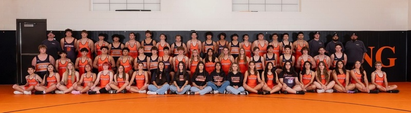 A large group of male and female teens in orange wrestling uniforms sit and stand in a gym while posing for a photo.