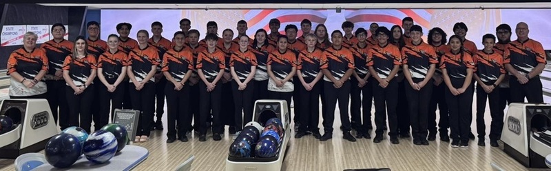 A large group of male and female teens in orange, black and white bowling shirts pose with their male and female coaches behind three bowling ball returns full of bowling balls.