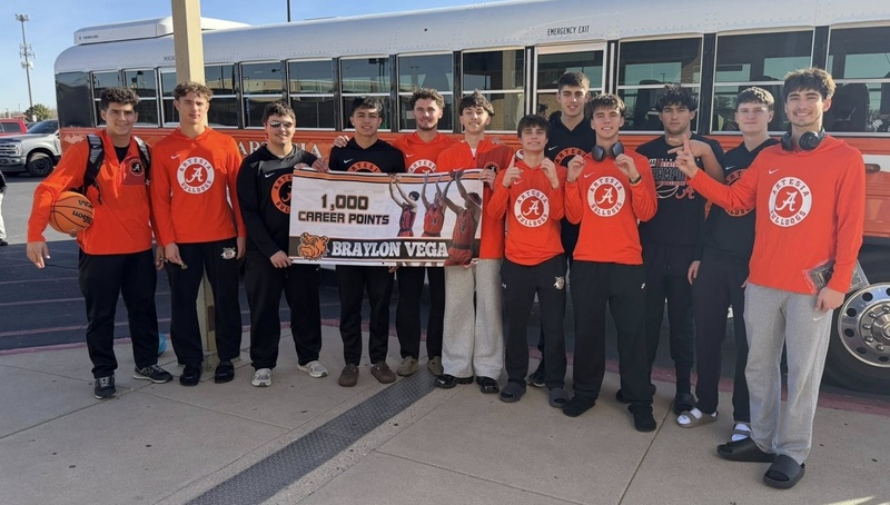 A group of teen boys in orange and black clothing pose for a photo outside an orange and white bus holding a banner celebrating Braylon Vega's 1,000th career point.