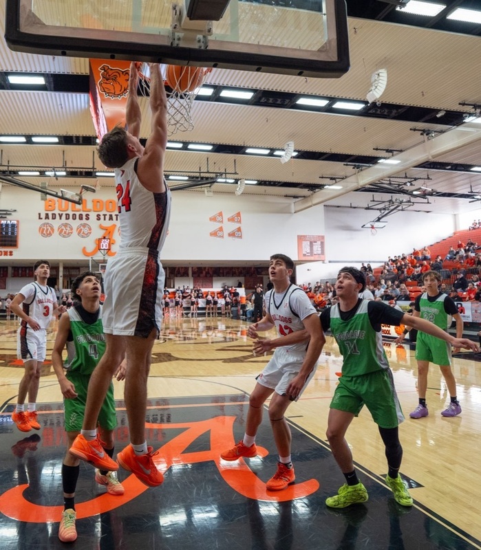 A teen boy in a white and orange basketball uniform is seen from the side dunking the ball as teammates and opponents in green look on.