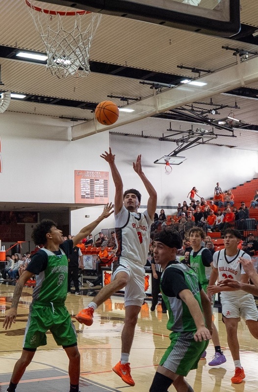 A teen boy in a white and orange basketball uniform leaps into the lane while shooting the ball as two opponents in green attempt to defend.