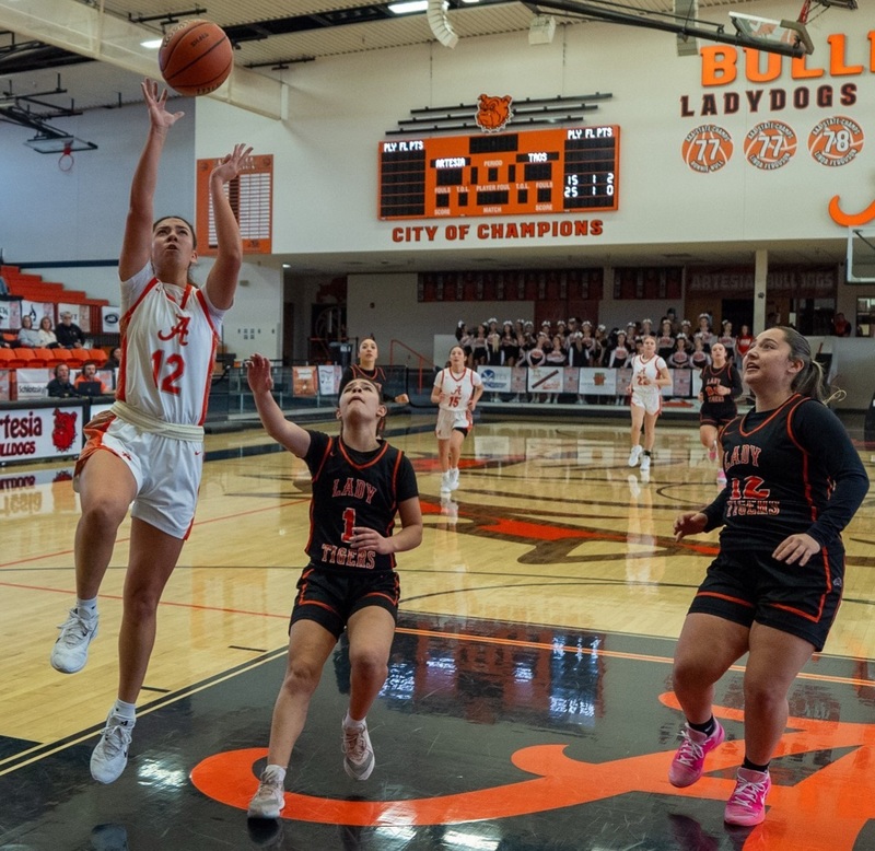 A teen girl in a white and orange basketball uniform goes in for a right-handed layup ahead of two opponents in black and orange.