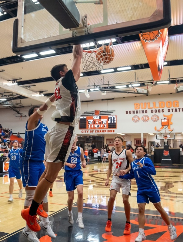 A teen boy in a white, orange and black basketball uniform hangs from the rim by his right hand after dunking the ball as teammates and opponents in blue look on.