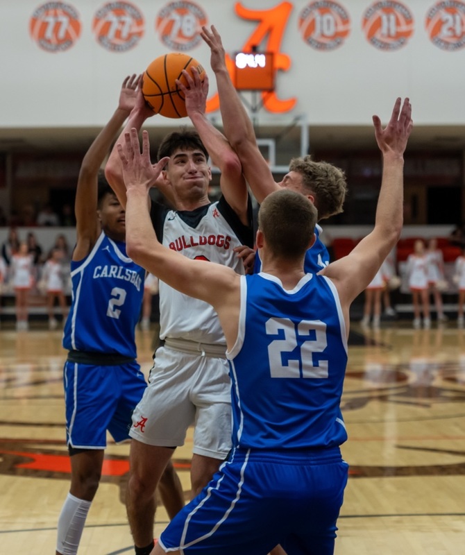 A teen boy in a white, orange and black basketball uniform holds the ball above his head with both hands as he's surrounded by opponents in blue.