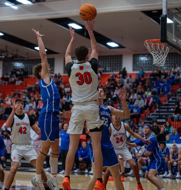 A teen boy in a white, orange and black basketball uniform is seen from behind as he jumps and releases the ball with his right hand as opponents in blue attempt to defend.