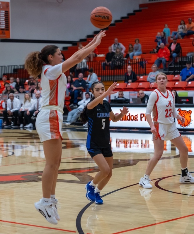 A teen girl in a white and orange basketball uniform jumps from behind the three-point arc as she releases the ball. A teammate and an opponent in black and blue look on.