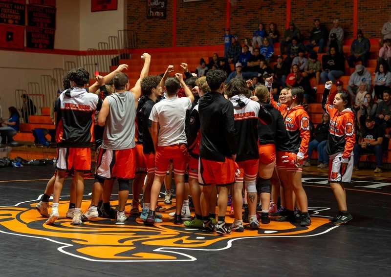 A large group of male and female teens in shirts and shorts gather in the middle of a black mat with an orange Bulldog logo and raise their hands as they break the huddle.