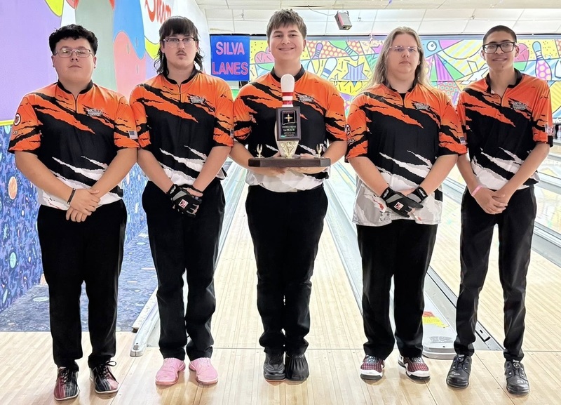 Five male teens in orange, black and white bowling shirts pose for a photo at a bowling alley. The center teen holds a trophy in the shape of a bowling pin.