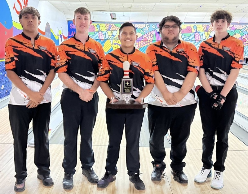 Five male teens in orange, black and white bowling shirts pose for a photo at a bowling alley. The center teen holds a trophy in the shape of a bowling pin.